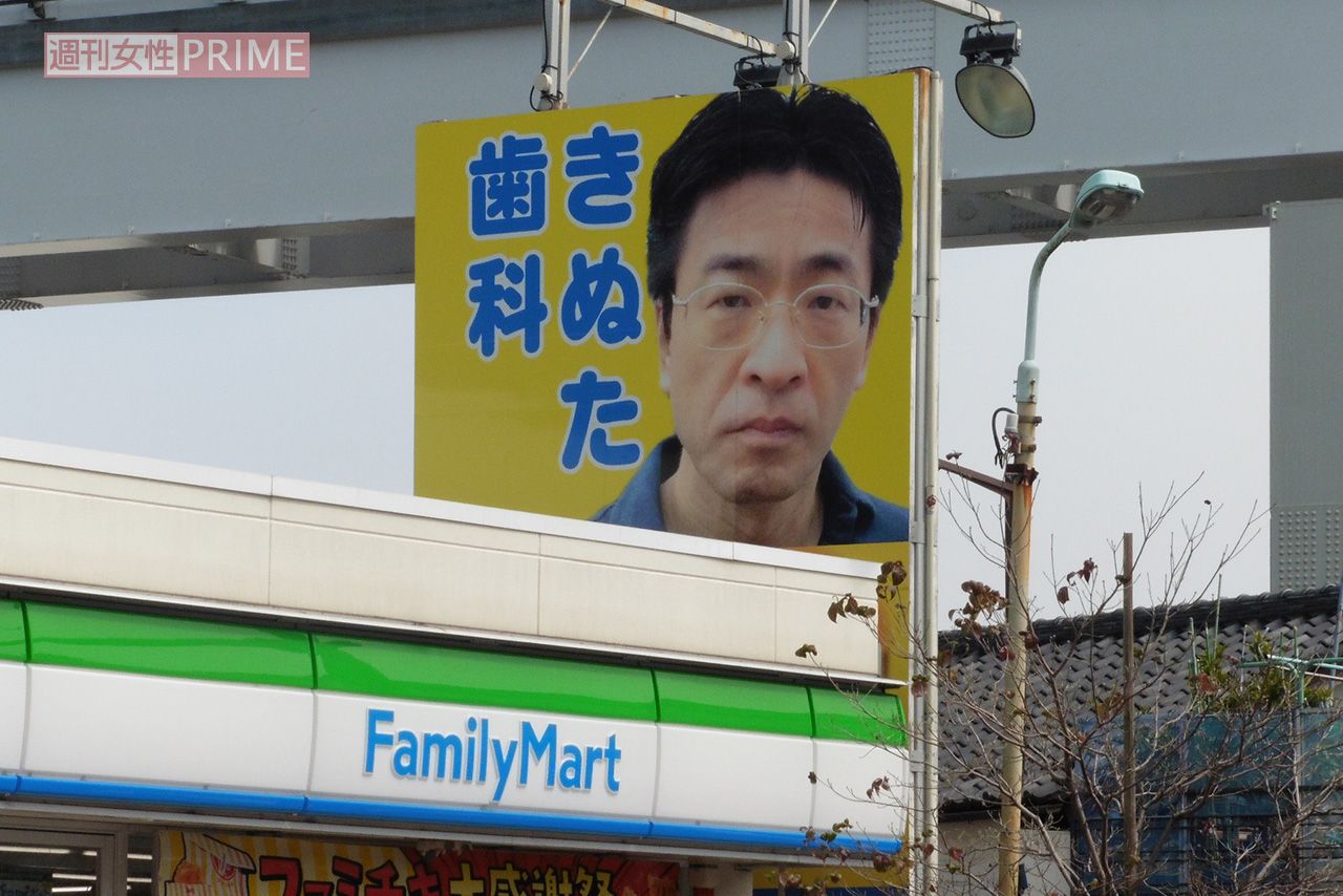 『きぬた歯科』看板マニアが260箇所から選ぶ「オススメ看板3選」その2「川崎街道と多摩都市モノレールが交わる箇所、ファミリーマート日野高幡店前にあります。当初は笑顔の院長の写真でしたが、ファミリーマートに阻まれた関係でデザイン変更を余儀なくされることに。結局怪訝な表情の院長が張り重ねられ、今に至ります」（『看板研究家』D.J.マメ氏）