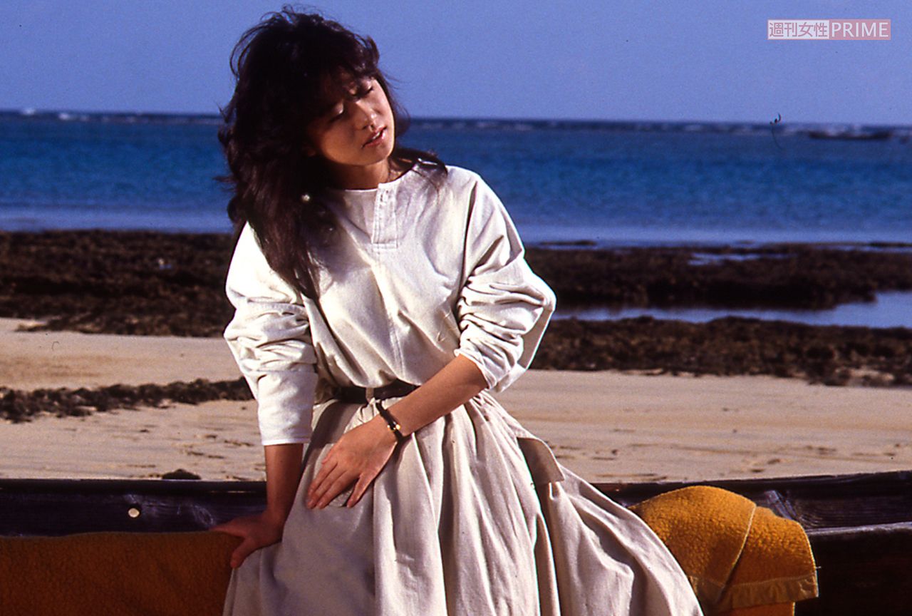 海辺でたそがれる中森明菜。青色の背景に白色のトップスがよく映える